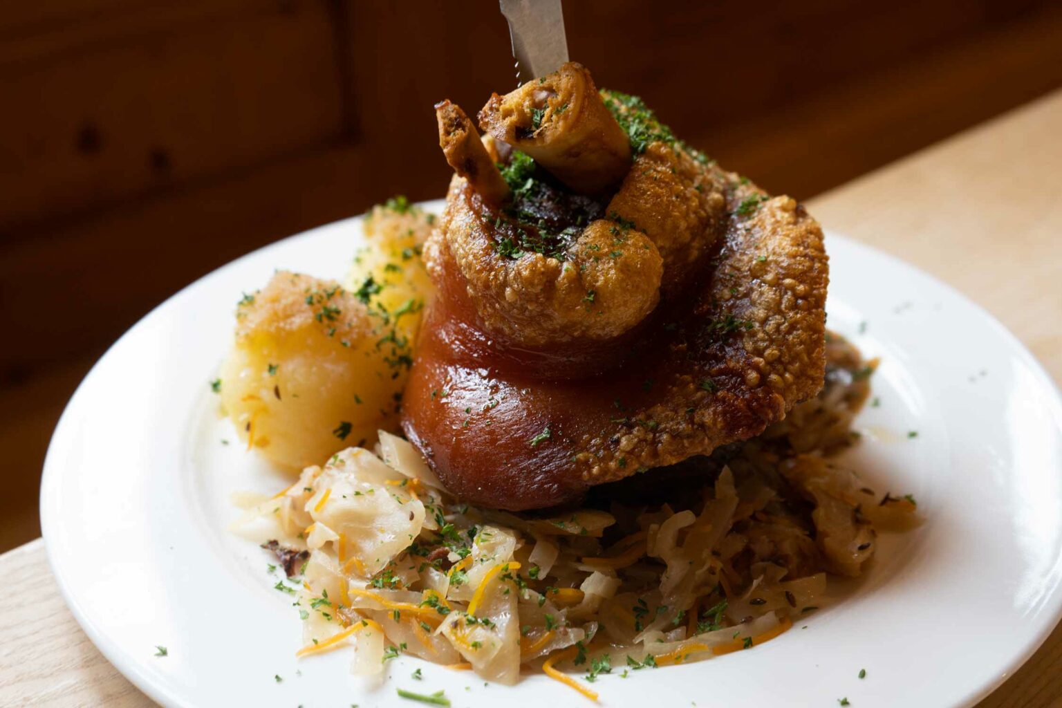 Knusprige Schweinshaxe auf Sauerkraut mit Kartoffelbeilage und Kräutern angerichtet.