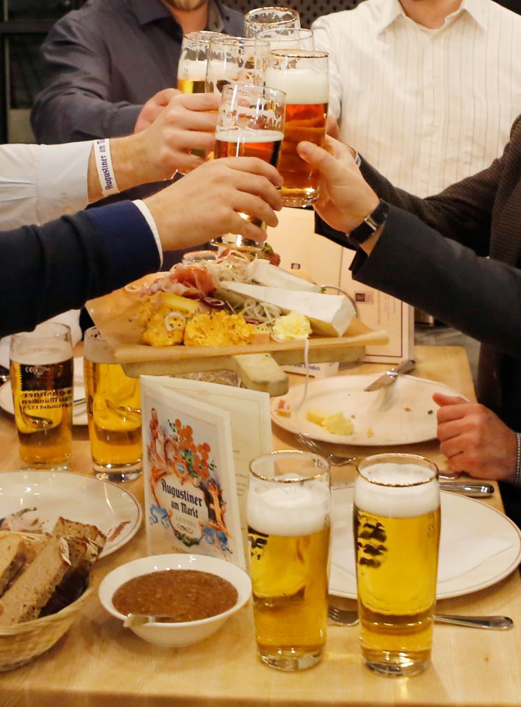 Mehrere Gäste stoßen mit frisch gezapften Augustiner-Bieren an, während eine reichhaltige bayerische Brotzeit auf dem Tisch serviert wird.