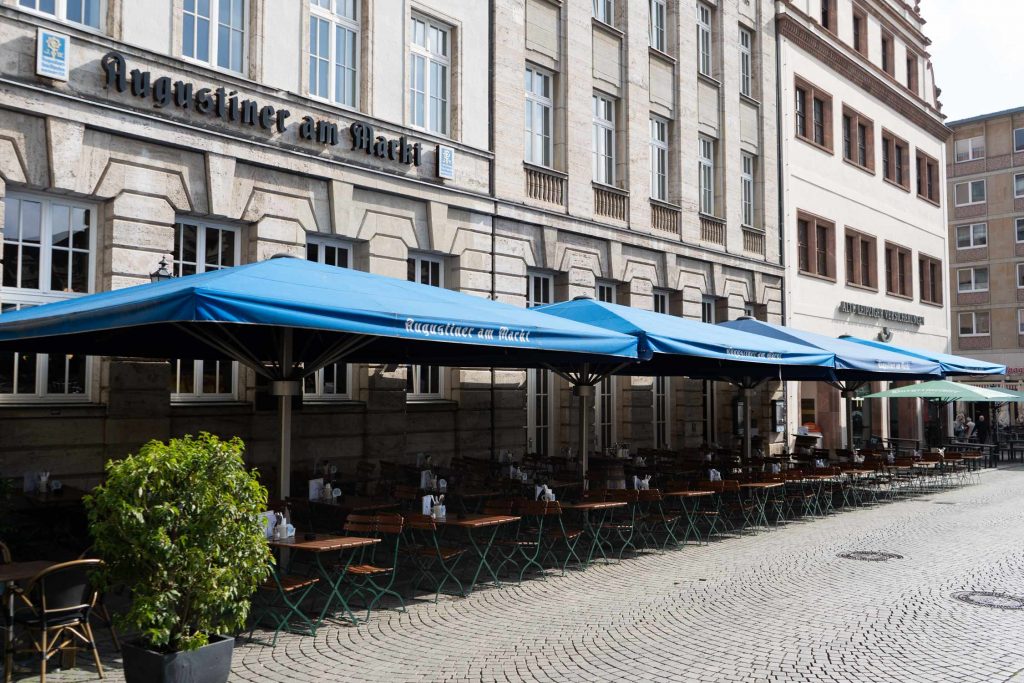 Der großzügige Außenbereich mit blauen Augustiner-Markisen erstreckt sich entlang der belebten Innenstadtstraße.