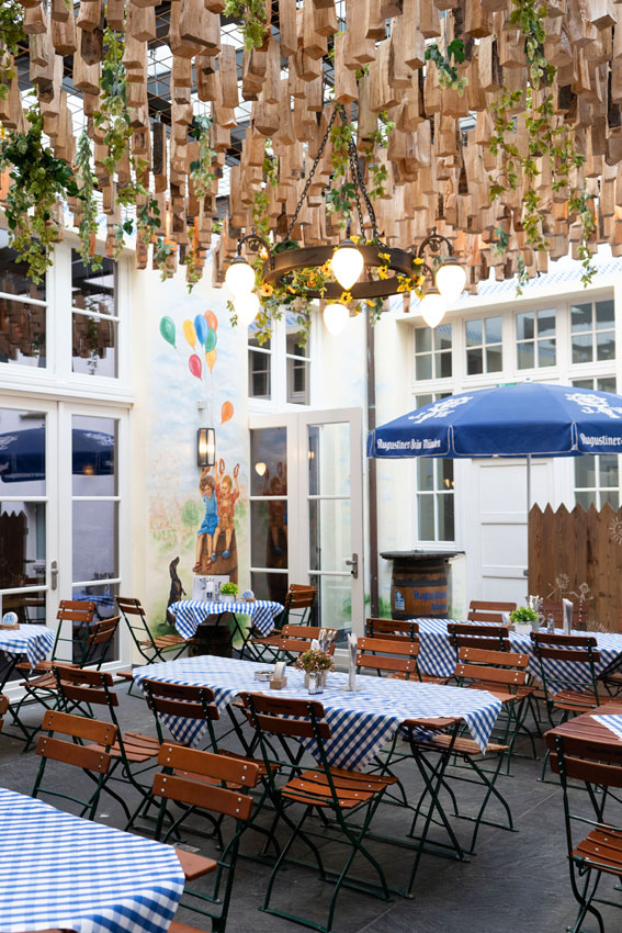 Heller Innenhof-Biergarten mit blau-weiß gedeckten Tischen, rustikalen Holzstühlen und dekorativer Hopfenbegrünung schafft eine gemütliche Atmosphäre.