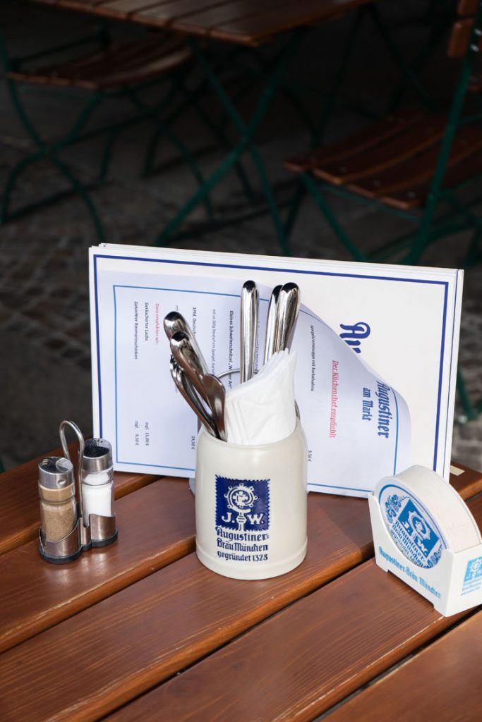Speisekarte, Besteck, Gewürze und ein Augustiner-Gefäß sind sorgfältig auf einem rustikalen Holztisch arrangiert.