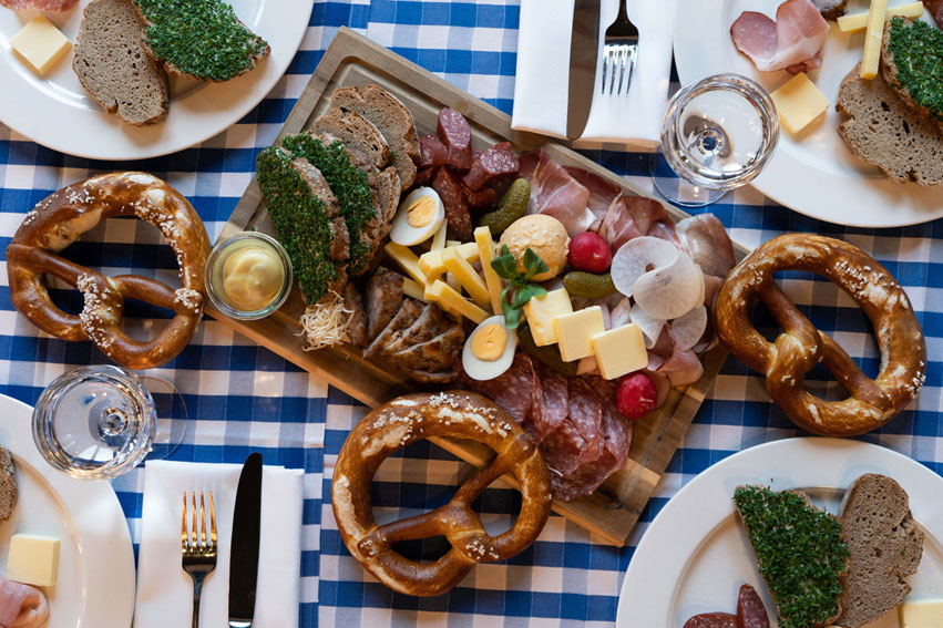 Eine reichhaltige bayerische Brotzeit mit Wurst, Schinken, Käse, Eiern, Brot und Brezeln ist auf einem Holzbrett angerichtet.